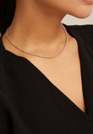 Primer plano de una persona que lleva un collar de cuentas plateadas y una blusa negra de escote en V, mostrando parte del cuello y la parte inferior de la cara.