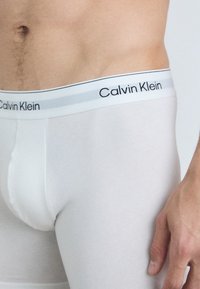 Boxeri albi Calvin Klein dintr-un material moale și elastic. Au un brâu contrastant alb și gri cu logo-ul mărcii.