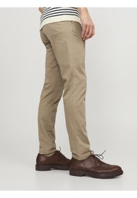 Uomo che indossa pantaloni chinos beige e scarpe stringate in pelle marrone scuro, in piedi contro uno sfondo leggero e neutro.