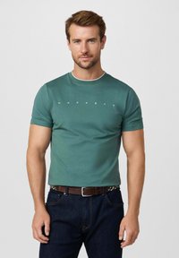 Camiseta de color verde oscuro con cuello redondo y mangas cortas. Presenta letras blancas "HACKETT" en el pecho. Se lleva con un cinturón marrón.