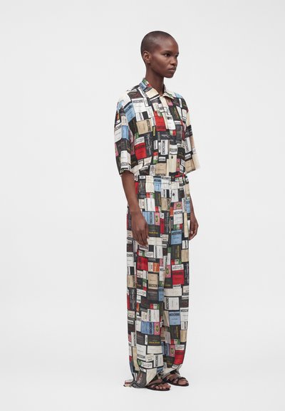 Femme aux cheveux courts portant une combinaison colorée et ample recouverte de divers patchs du logo Moschino, debout devant un fond uni.