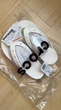 Weiße Crocs mit strukturiertem Design, aus leichtem Material, verpackt in transparenter Plastikfolie. Enthält Etiketten mit Größen- und Pflegeinformationen.