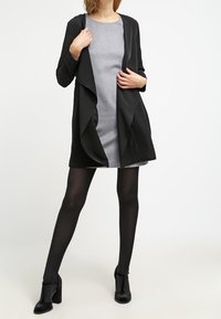 Svart öppet kort över en grå klänning, parat med svarta högklackade ankelstövlar. Outfiten har släta tyger och en smal passform.