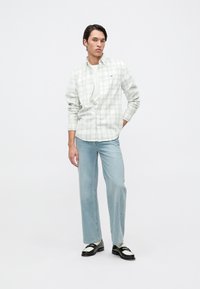 Cămașă cu nasturi,în carouri gri deschis, cu mâneci lungi, purtată deschisă peste un tricou alb. Blugi drept, de culoare albastră deschisă, și mocasini negri cu accente albe.