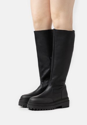 Alti stivali neri in pelle con suole spesse, indossati su gambe nude, mostrati su uno sfondo bianco.