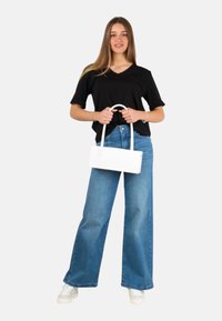 Borsa bianca rettangolare con manico superiore, tenuta da una persona che indossa una t-shirt nera a V e jeans larghi blu. Sneakers bianche visibili.