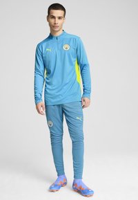 Lichtblauw sportief trainingspak met ritskraag, voorzien van neongele accenten. Inclusief logo-opdracht en een aansluitend ontwerp. Bijpassende kleurrijke sportschoenen.