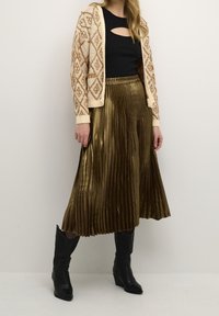 Stickad cardigan med diamantmönster i creamfärg och rost, i kombination med en metallic guld veckad kjol och svarta cowboystövlar.