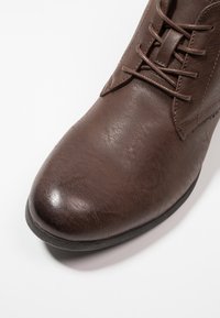 Bota de cuero marrón con acabado texturizado, diseño con cordones, punta redondeada y un ligero tacón. Presenta acentos cosidos en los lados.