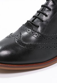 Chaussure en cuir noir avec des détails en brogue, texture lisse, bout arrondi et semelle en caoutchouc marron légèrement brillante.