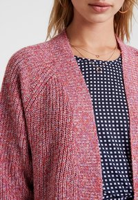 Cardigan lavorato a maglia rosa e arancione con apertura frontale, tessitura testurizzata e vestibilità ampia, indossato sopra una camicetta a quadri blu navy con quadrati bianchi.