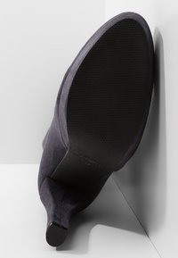 Scarpa con tacco alta in suede viola scuro vista dal basso, con una suola in gomma nera testurizzata e un design a punta.