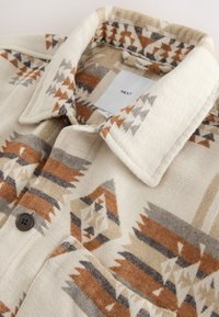 Chemise boutonnée en tissu tissé présentant un motif géométrique dans des nuances de beige, rouille et gris, avec un col classique et une étiquette.