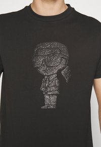 Camiseta negra con un gráfico metálico plateado texturizado de un personaje de dibujos animados con gafas, vestido con traje y corbata, con las manos entrelazadas.