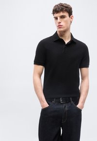 Νεαρός άνδρας με κοντά σγουρά μαλλιά φοράει μαύρο κοντομάνικο πόλο με υφή και σκουρόχρωμα τζιν, στέκεται με τα χέρια στις τσέπες μπροστά σε απλό φόντο.