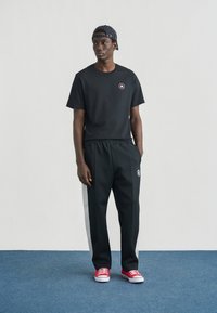 Μαύρο βαμβακερό μπλουζάκι με λογότυπο, σε συνδυασμό με μαύρο παντελόνι που έχει γκρι ρίγες στο πλάι. Το μοντέλο φοράει κόκκινα αθλητικά παπούτσια και ριγωτό καπέλο.