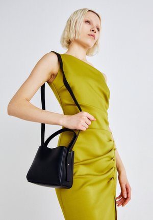 Mujer con vestido sin mangas color verde oliva sosteniendo un bolso negro con asas para el hombro y la mano sobre un fondo liso.