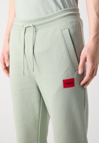 Pantalones de chándal verde claro hechos de un tejido suave con cintura elástica, que cuentan con bolsillos laterales y un parche del logo rojo "HUGO".