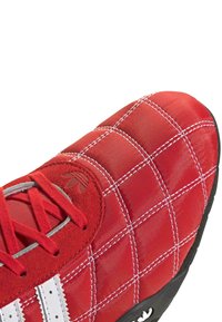 Röd sneaker med quiltad textur, vita detaljer och grå snören. Har en svart gummisula och en logotyp på tungan.