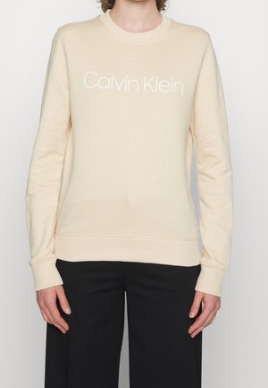 Persona che indossa un maglione beige a maniche lunghe con il testo "Calvin Klein" sulla parte anteriore e pantaloni neri.