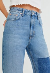 Jean en denim bleu clair taille haute avec une texture délavée et un patch de denim plus foncé sur la cuisse droite. Présente des éléments métalliques en cuivre et cinq poches.