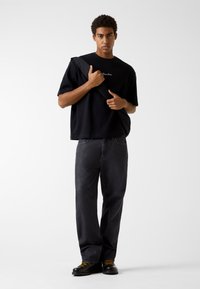 Czarny, oversize'owy t-shirt z krótkim rękawem i haftowanym logo, zestawiony z ciemnymi jeansami o prostych nogawkach oraz czarnymi sznurowanymi botkami z żółtymi sznurówkami.