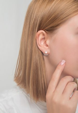 Femme aux cheveux blonds raides portant une boucle d'oreille argentée ronde avec un prolongateur ondulé en argent, touchant son visage avec un doigt manucuré.