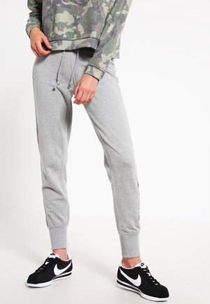 Person trägt graue Jogginghose mit Kordelzug und schwarze Nike-Sneaker mit weißem Swoosh, steht vor weißem Hintergrund.