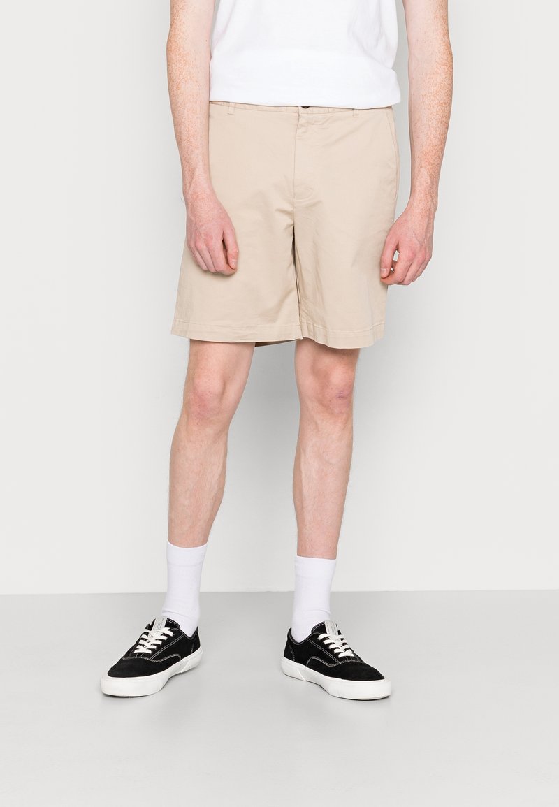 Shorts en coton beige au style classique, avec une taille plate, des poches latérales et un ourlet simple ; portés avec des baskets noires et des chaussettes blanches.