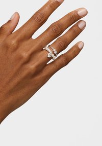 Ręka z dwoma srebrnymi pierścionkami na środkowym palcu, jeden z małymi perłami i zawieszkami w kształcie serca, na jednolitym jasnym tle.