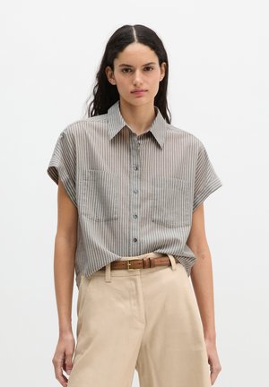 Jeune femme aux cheveux foncés porte une chemise rayée à manches courtes boutonnée, rentrée dans un pantalon beige avec une ceinture marron.