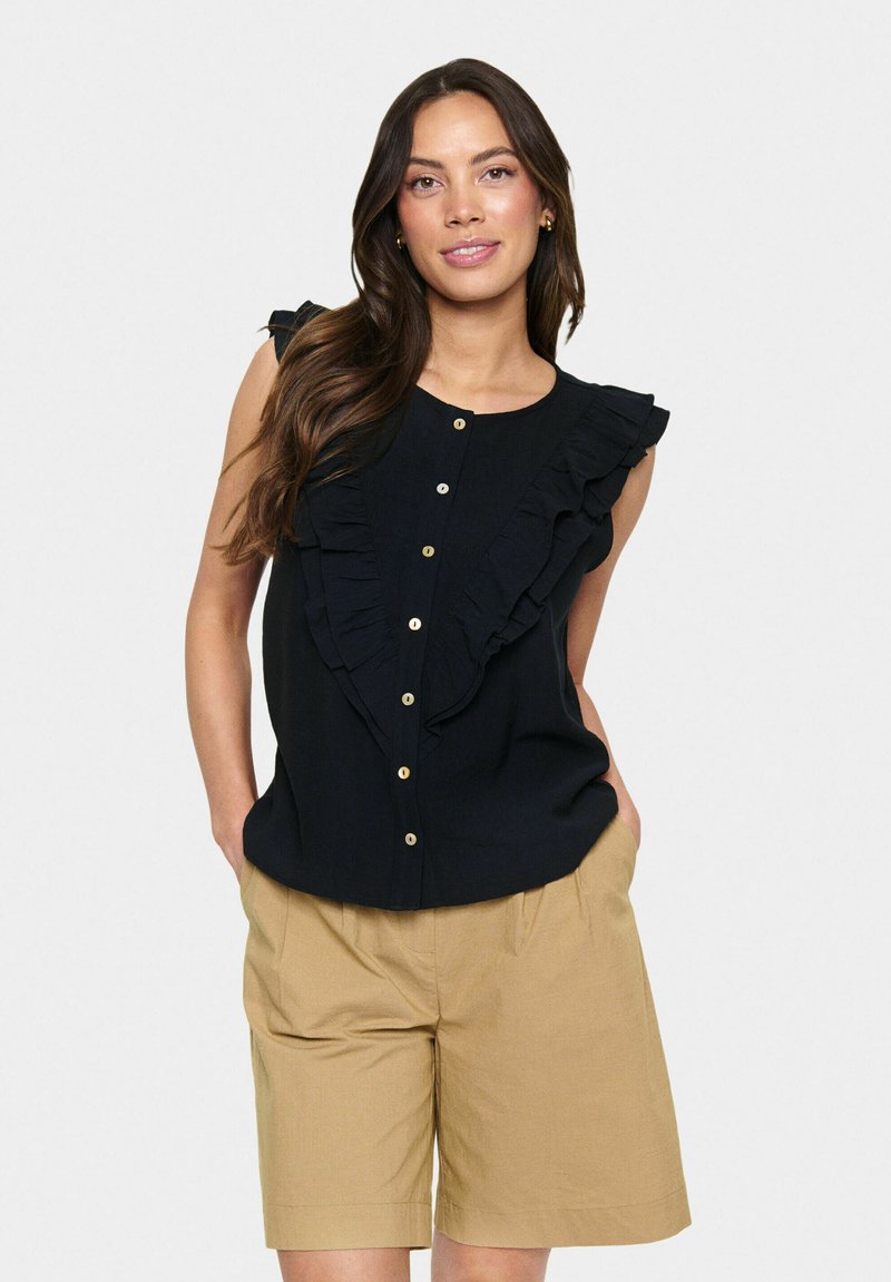 Blusa nera senza maniche con una parte anteriore arricciata, chiusura con bottoni e una leggera lucentezza, abbinata a pantaloni corti beige a vita alta.