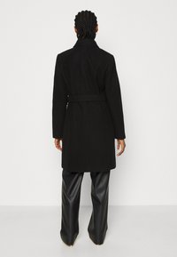 Abrigo negro de tela suave con cintura ceñida, mangas largas y un cuello clásico; combinado con pantalones anchos de cuero sintético negro.