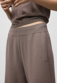 Close-up van een persoon in taupe gebreide broek met elastische tailleband en bijpassende mouwloze top, handen voorzichtig dichtbij de taille.