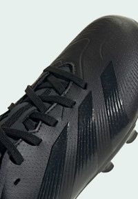 Botas de fútbol negras con una superficie texturizada que presenta puntos elevados y acentos angulares. Los cordones son negros y el diseño es aerodinámico.