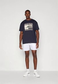 Μπλε μπλουζάκι με εκτυπωμένο γραφικό που απεικονίζει το «Monte Carlo» και έναν τενίστα, σε συνδυασμό με λευκά σορτς και λευκά αθλητικά παπούτσια.