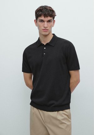 Młody mężczyzna o kręconych włosach stoi zwrócony przodem, ubrany w czarną krótką koszulkę polo i beżowe spodnie na jednolitym tle.