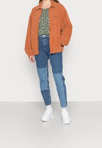 Oranžová manšestrová bunda přes zelený květinový top, spárovaná s patchworkovými modrými džíny. Bílí tenisky doplňují outfit na neutrálním pozadí.
