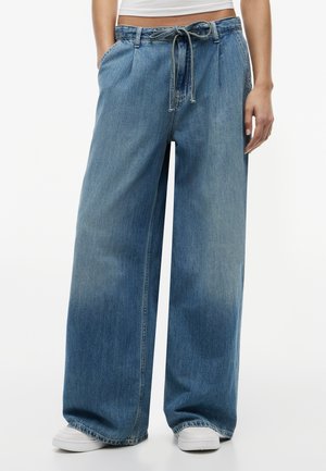 Kvinne iført løstsittende, vide blå jeans med snor i midjen og hvite joggesko, stående mot en ensfarget bakgrunn.