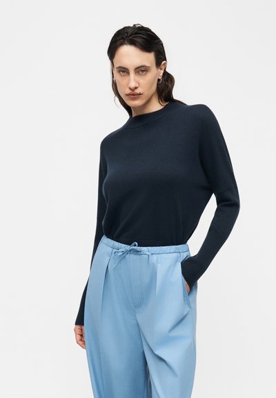 Une femme aux cheveux foncés porte un pull ajusté noir à manches longues et un pantalon ample bleu clair avec une taille à cordon, posant avec une main dans la poche.