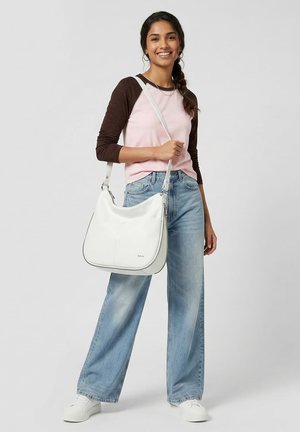 Frau trägt ein rosa-braunes Langarmshirt und weite Jeans, hält eine weiße Schultertasche und lächelt vor einem schlichten Hintergrund.