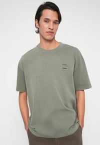 Ung man med mörkt hår som bär en lös olivgrön t-shirt med "SAMSØE"-logotypen, står med händerna i fickorna framför en enfärgad vit bakgrund.