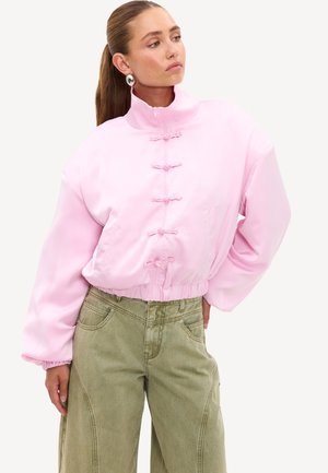 Femme aux longs cheveux attachés portant une veste rose clair oversize avec des boutons grenouille et un pantalon large vert olive, posant la main sur la hanche.