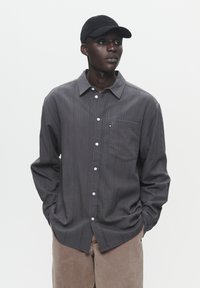 Gray striped long-sleeve shirt with a button-down collar and front pocket, paired with beige pants and a black cap.