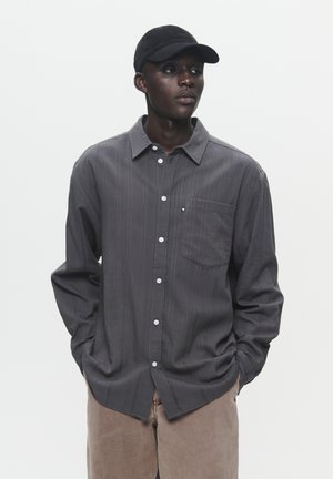 Gray striped long-sleeve shirt with a button-down collar and front pocket, paired with beige pants and a black cap.