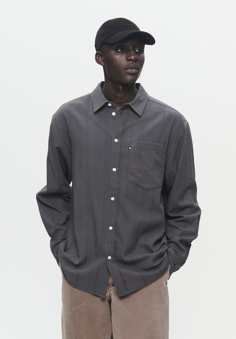 Gray striped long-sleeve shirt with a button-down collar and front pocket, paired with beige pants and a black cap.