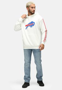 Weißer Kapuzenpullover mit einem blauen und roten Büffel-Design, gestreiften roten Akzenten an den Ärmeln und einer vorderen Kängurutasche. Kombinieren Sie ihn mit hellen Jeans.