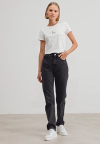 Jeans negros de tiro alto con piernas rectas, combinados con una camiseta blanca ajustada que presenta un pequeño logo gris. Zapatillas blancas completan el look.
