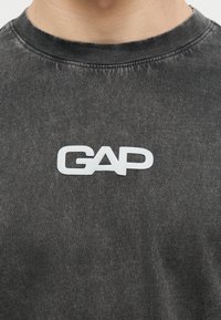 Κοντινό πλάνο ενός ατόμου που φοράει μια ξεβαμμένη μαύρη μπλούζα με λευκό λογότυπο GAP τυπωμένο στο στήθος.