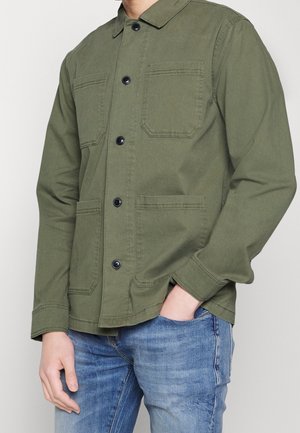 Mężczyzna w oliwkowo-zielonej kurtce koszulowej z czterema przednimi kieszeniami, zestawionej z niebieskimi dżinsami, pozujący z jedną ręką w kieszeni.